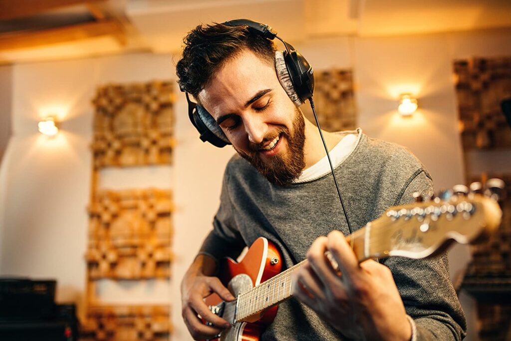 A warmly-lit photograph of a good looking man with a well kept beard, wearing a pair of DT770 headphones and playing electric guitar.